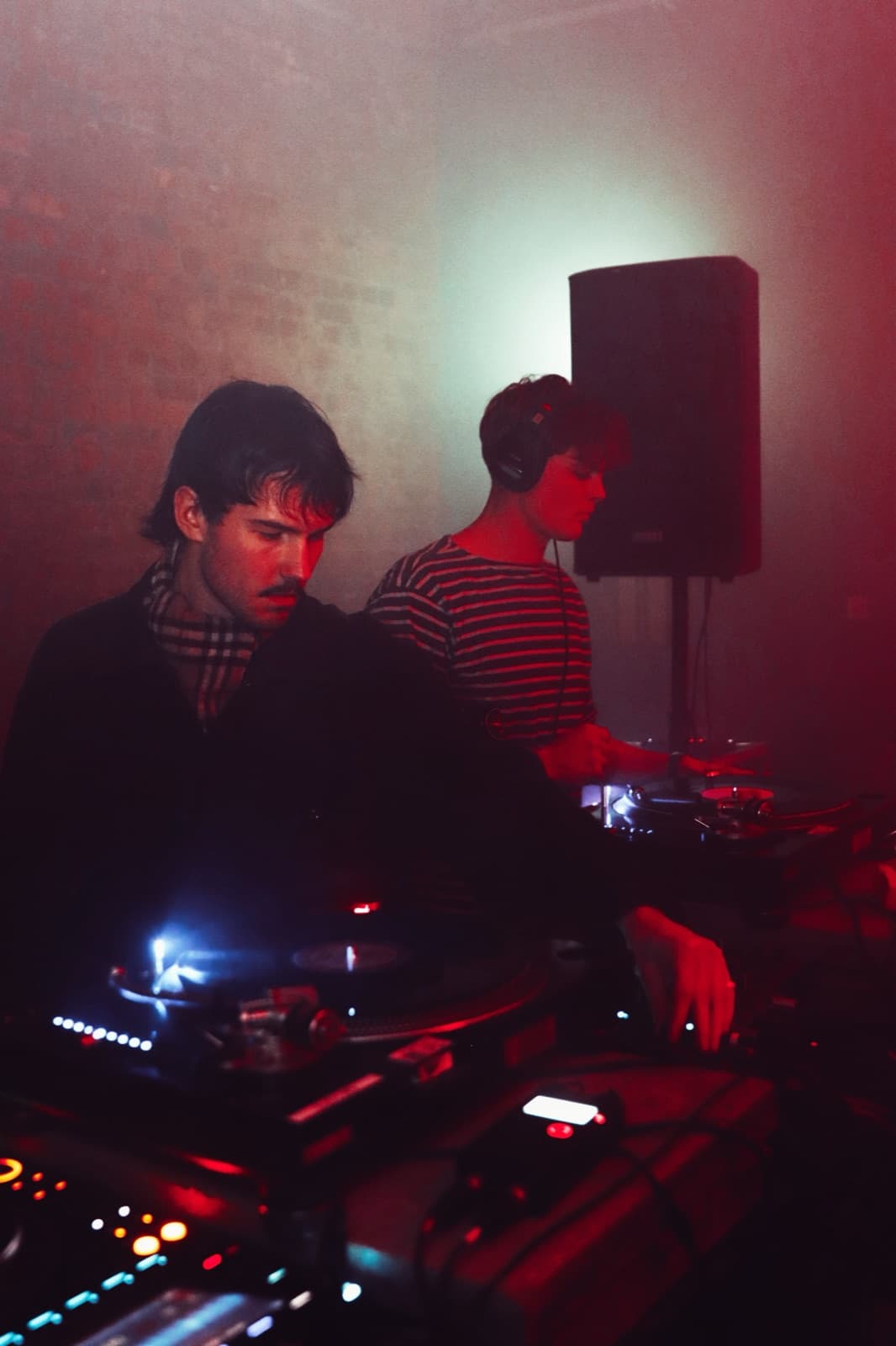 Two DJs working the turntables, one in headphones and a striped top, in red light