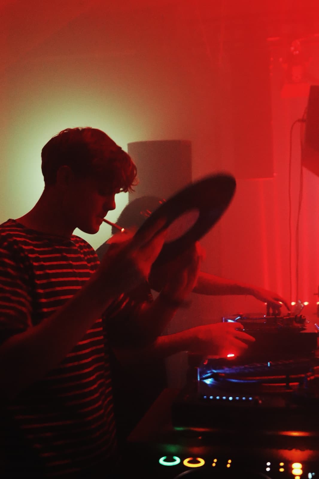 DJ lifting a vinyl record from the deck with a cigarette, red light behind